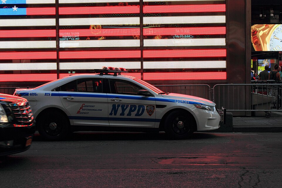 NYPD patrol car in Times Square in front of an illuminated American flag display.