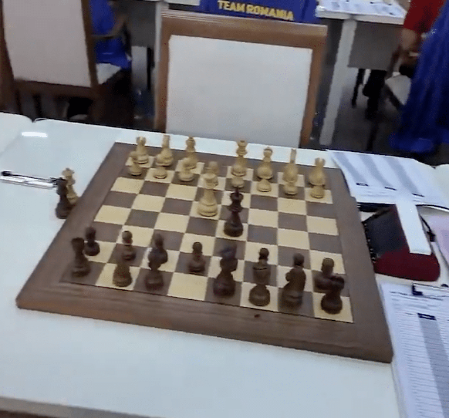 Chessboard with wooden pieces on a tournament table beside an empty chair.