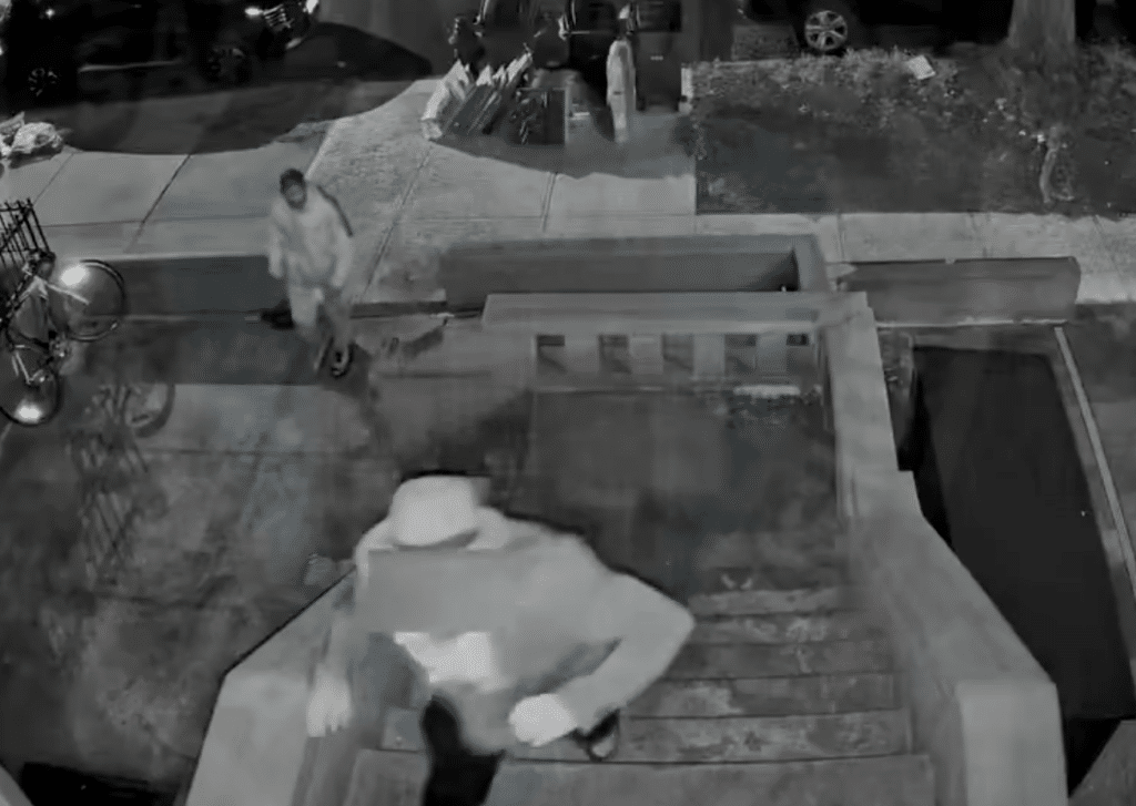 Black-and-white surveillance image showing one person on the steps of a house and another approaching from the sidewalk at night.
