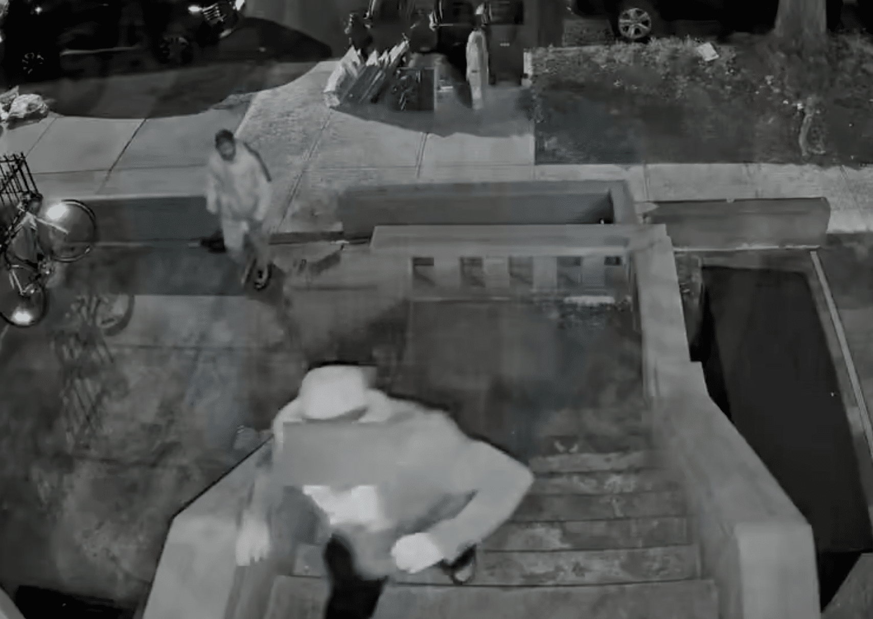Black-and-white surveillance image showing one person on the steps of a house and another approaching from the sidewalk at night.