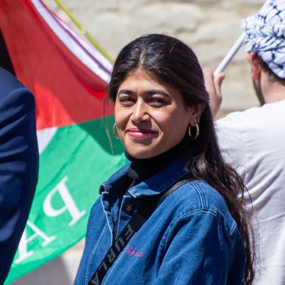 Rima Hassan wearing a denim jacket, with a Palestinian flag behind her.