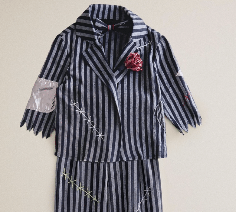 Zara children’s costume with gray-and-blue vertical stripes, crisscross stitching, ragged sleeves, and a red lapel flower.