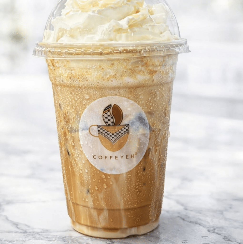 Cold coffee drink topped with whipped cream in a clear cup bearing Coffeyeh’s keffiyeh-themed logo.
