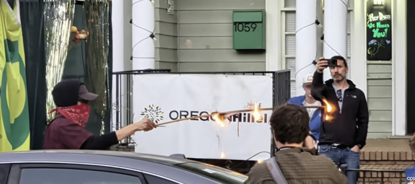 Masked protester burns an American flag outside Oregon Hillel in Eugene as a bystander records with a phone.